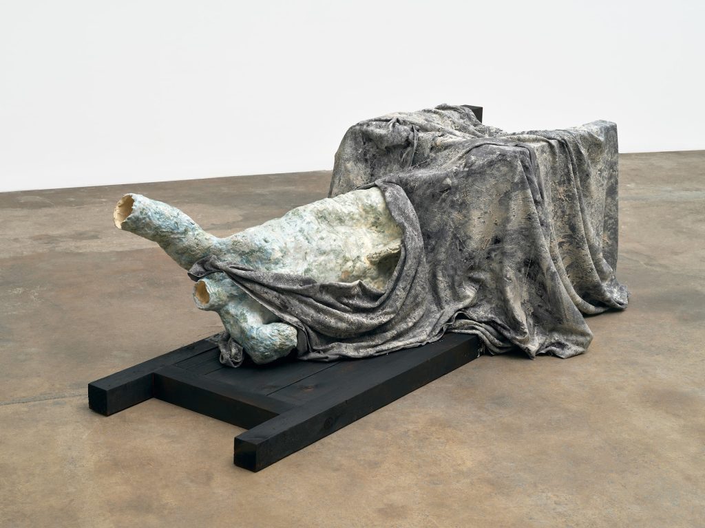 This image depicts an artwork by Liz Glynn titled "Toppled Zeus." This artwork was created in 2023 and measures 30" x 87" x 46" [HxWxD] (76.2 x 220.98 x 116.84 cm). Its medium is Glazed ceramic torso, burnt wood chair, resin-fixed wool drapery.