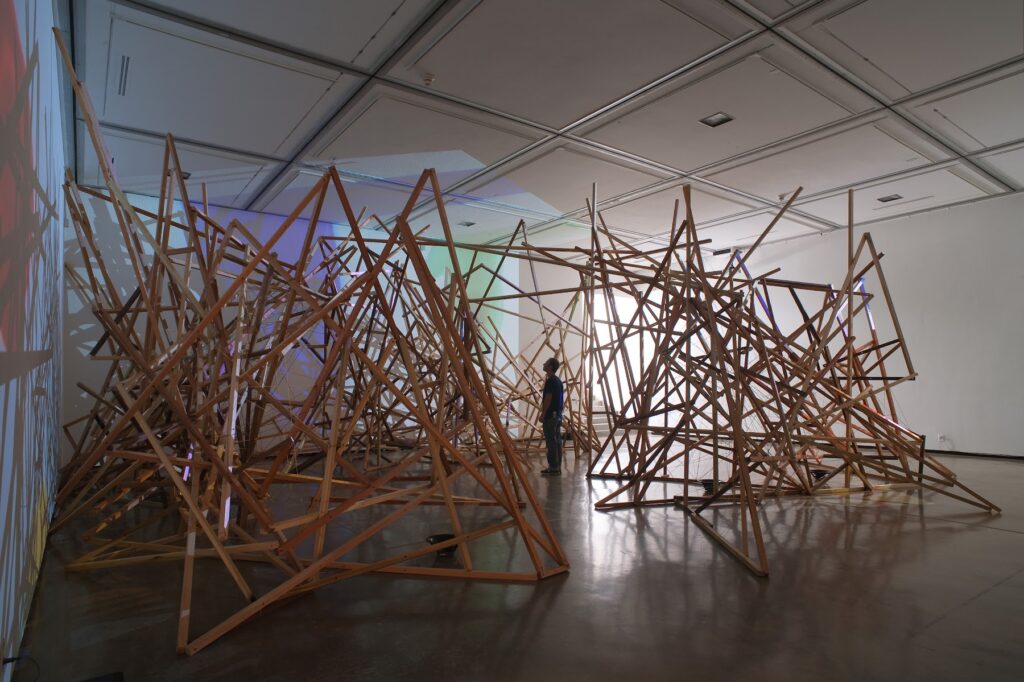 This image depicts an artwork by Steve Roden titled "bowrain." This artwork was created in 2010 and measures Site specific installation; Dimensions variable: 45'  x 45' [HxW] (1,371.6 x 1,371.6 cm). Its medium is Wood, wire, twine, sound composition, 6 audio speakers, 3 cd players, 3 stereo amplifiers, ink on 16mm film stock transferred to video, 3 dvd players, 3 video projectors.