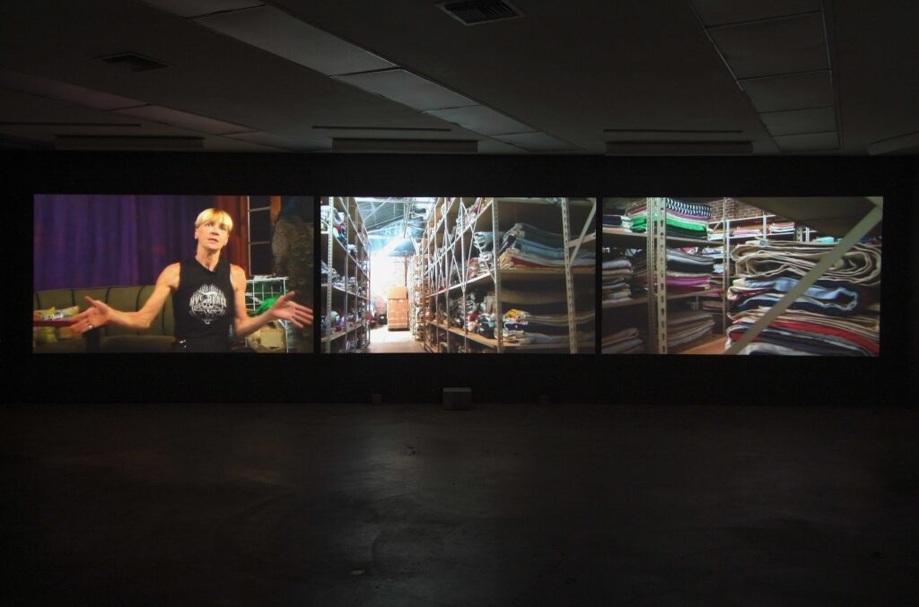 This image depicts an artwork by Andrea Bowers titled "The Weight of Relevance." This artwork was created in 2007 and measures TRT: 26:15 minutes with sound; projection 79" x 432"  [HxW] (200.66 x 1097.28 cm). Its medium is 3 channel video (color with sound) [3 dvds, 3 dvd players, 3 video projectors, speakers].