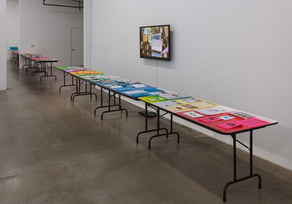 This image depicts an artwork by Andrea Bowers titled "Workers' Rights Table." This artwork was created in 2012 and measures Dimensions variable, 18" x 15" x 8" (Book). Its medium is "Labor is Entitled to All It Creates" (Political Flyers, colby posters and calligraphy bound in book), folding work tables, political flyers and colored vinyl.