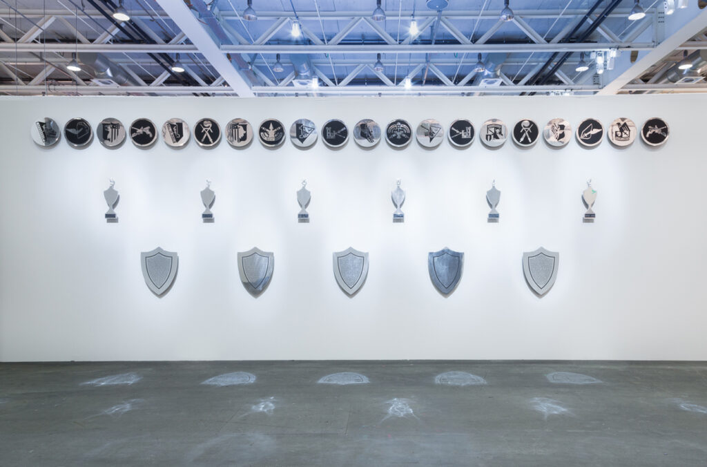 This image depicts an artwork by Mary Kelly titled "Gloria Patri." This artwork was created in 1992 and measures 31 Units Total. Installation Dimensions variable: 5 Shields: 29" x 24" x 2 ¹⁄₂" [HxWxD] (73.66 x 60.96 x 6.35 cm) each, 6 Trophies: 24" x 8" x 2" [HxWxD] (60.96 x 20.32 x 5.08 cm) each, 20 Discs: 17" x 2 ¹⁄₄" [Diameter x Depth] (43.18 x 5.71 cm) each. Its medium is Etched and polished aluminum.