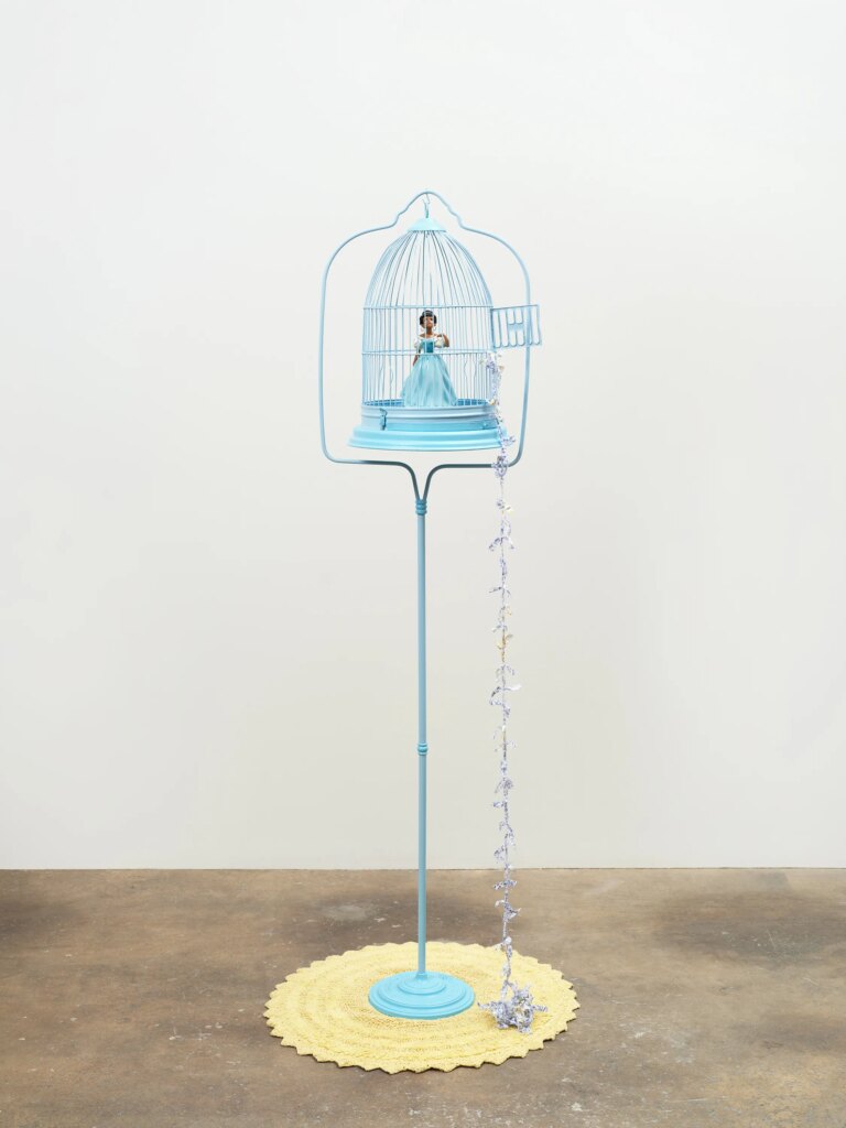 This image depicts an artwork by Genevieve Gaignard titled "Chasing the Moon." This artwork was created in 2022 and measures 67" x 15" x 15" [HxWxD] (170.18 x 38.1 x 38.1 cm). Its medium is Birdcage with stand, custom porcelain figurine, doily, fabric ladder.